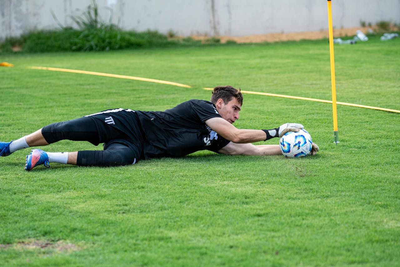A goalkeeper dives to catch a soccer ball during practice on a lush green field.